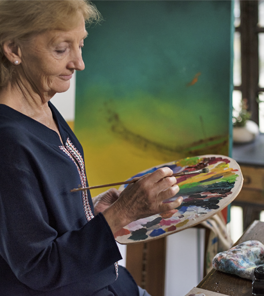 An older woman standing in front of a canvas with a painting on it. She is holding a pallet and a paint brushing looking lost in thought.