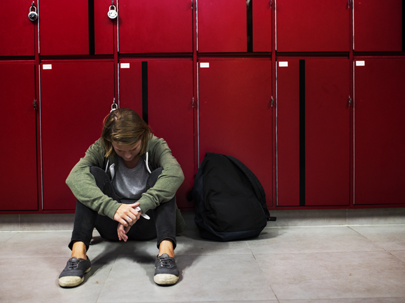 A young man sitting in a hallway next to locker in a school.