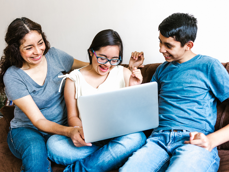 A family sitting on a couch looking at a laptop. The family is laughing and smiling.