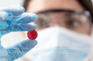 A stock image of a woman blurred out in the background with her hand in focus holding a covid-19 virus model.