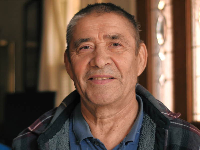 An older man smiling at the camera with light coming from a window in the background.