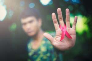 A blurred out man in the background with his hand in focus holding a red ribbon the represents HIV support.