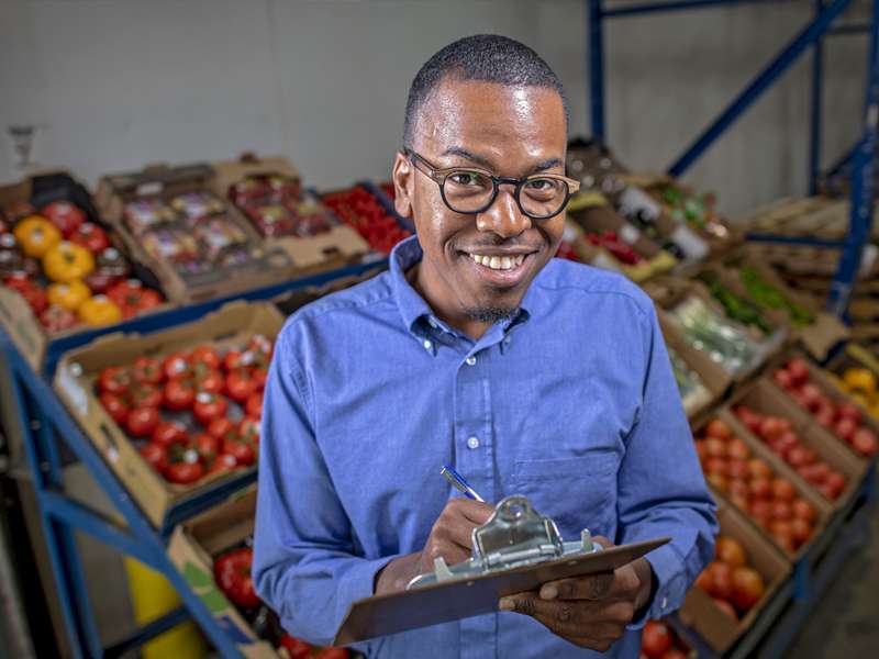 a young man holding a clipboard with vegetables behind him in boxes.