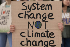 A cardboard protest sign held by a person reads, “System change not climate change,” with a small drawing of the Earth inside the letter O in the word “not.”